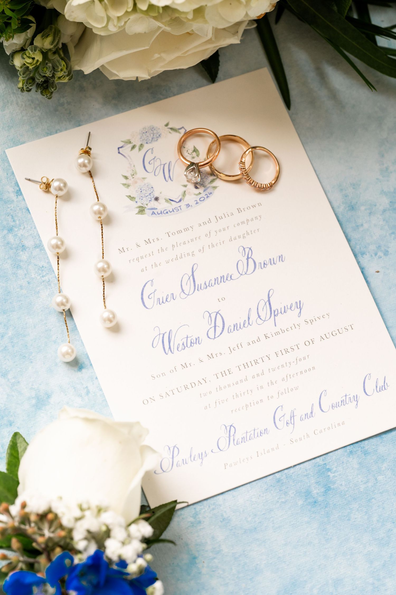 Elegant wedding invitation with gold rings and white floral details on a light blue background.