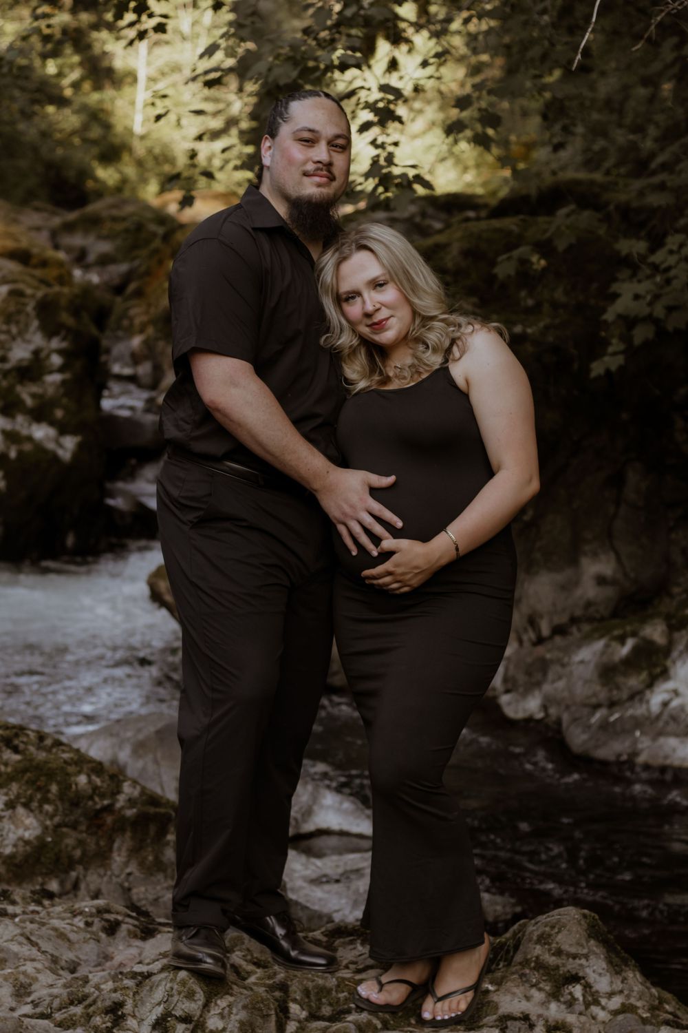Couple in matching dark outfits posing by deschutes river, woman in black dress cradling baby bump during maternity photo session.