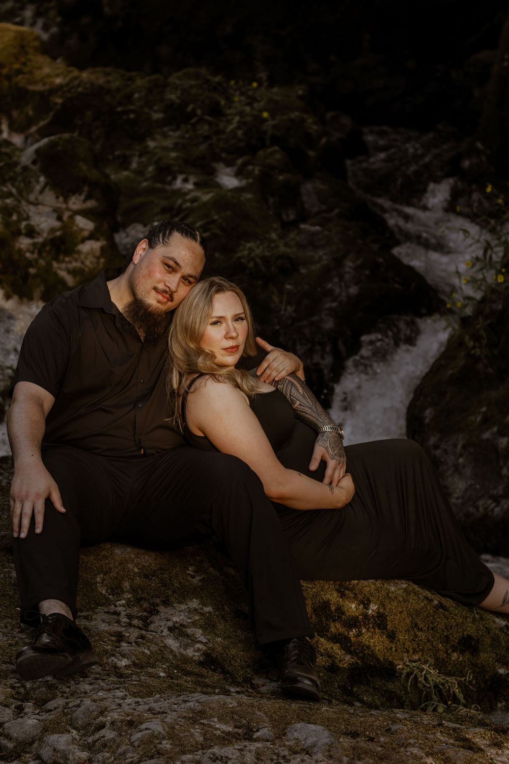 A couple in black outfits sits on rocks near deschutes river waterfall, the man resting his head on the woman's shoulder.