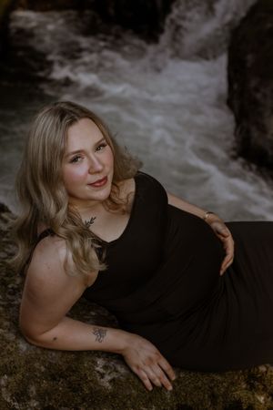 Blonde woman in black dress reclining on rocks near rushing deschutes river waterfall, smiling softly at camera.