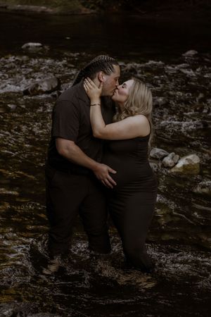 Couple standing and  kissing in deschutes river, woman in black dress showing baby bump, man in dark shirt, moody outdoor maternity photo.