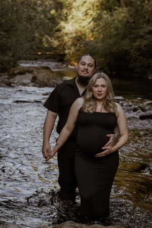 Couple maternity photo in deschutes river, woman in black dress cradling baby bump, man in black shirt standing behind her.