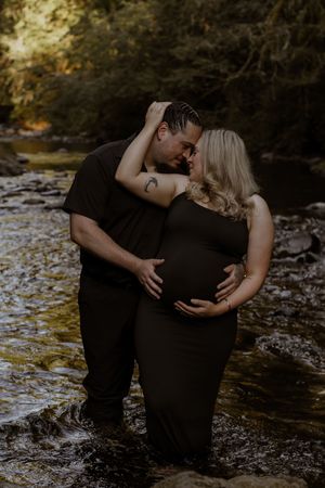 Couple in matching black outfits sharing a tender moment in deschutes river, woman in maternity dress cradling baby bump.