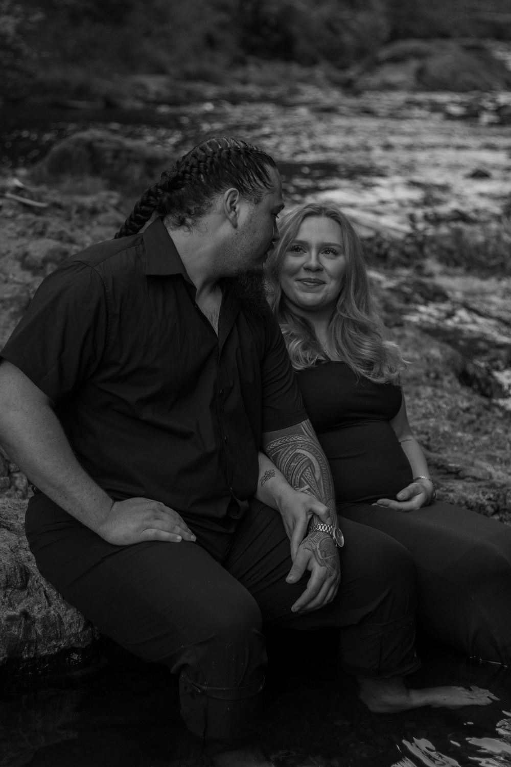 Black and white maternity photo of a couple sitting by a deschutes river, looking at eachother