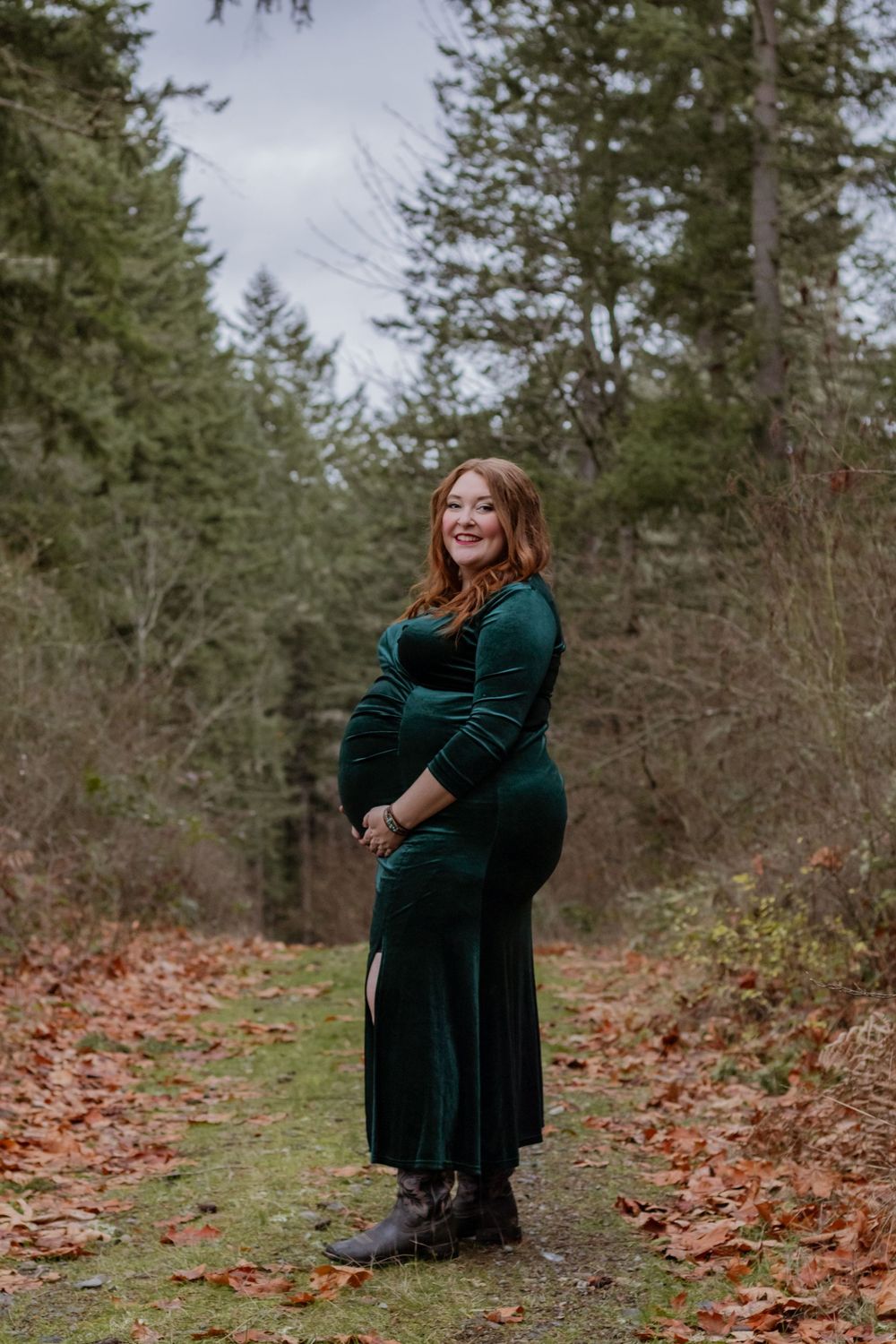 Happy pregnant woman in green velvet dress standing on leaf-covered forest path smiling.