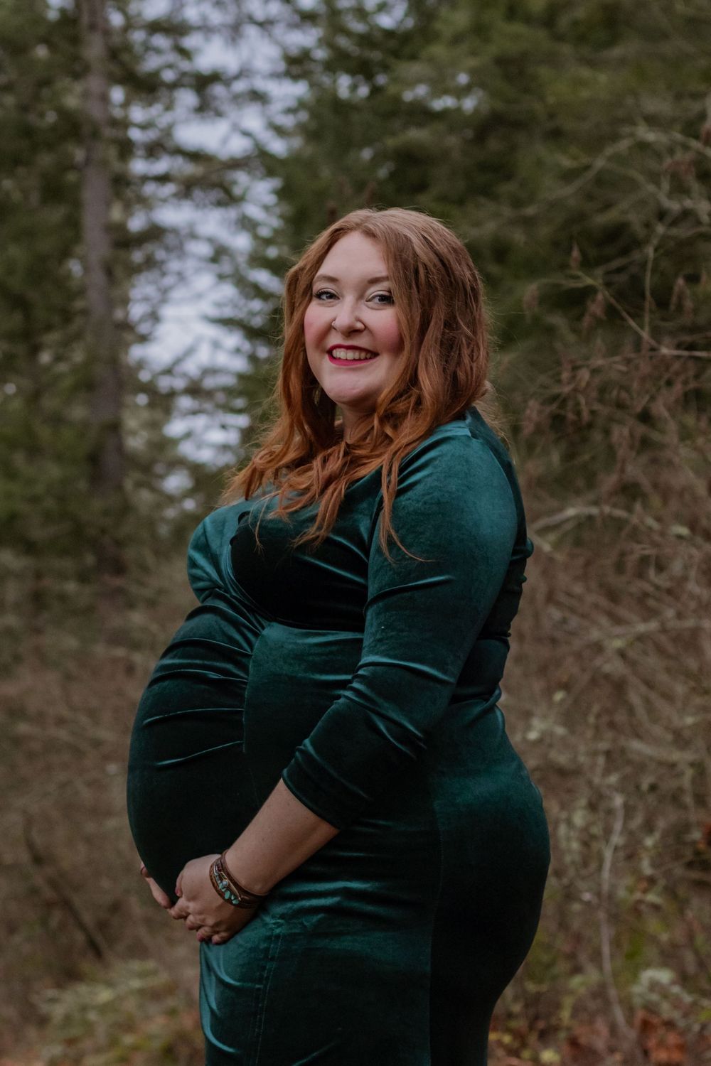 Smiling pregnant woman in emerald green velvet dress cradling baby bump in forest setting.