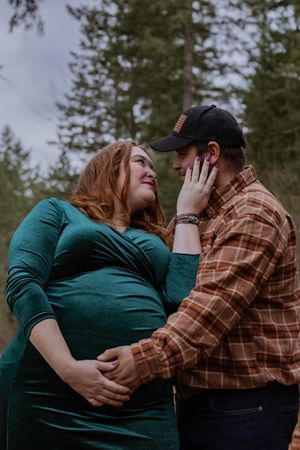 Pregnant couple sharing a tender moment in forest, woman in green dress touching mans face.