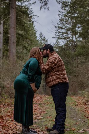 Pregnant couple sharing a kiss on a leaf-covered forest path surrounded by tall pine trees.