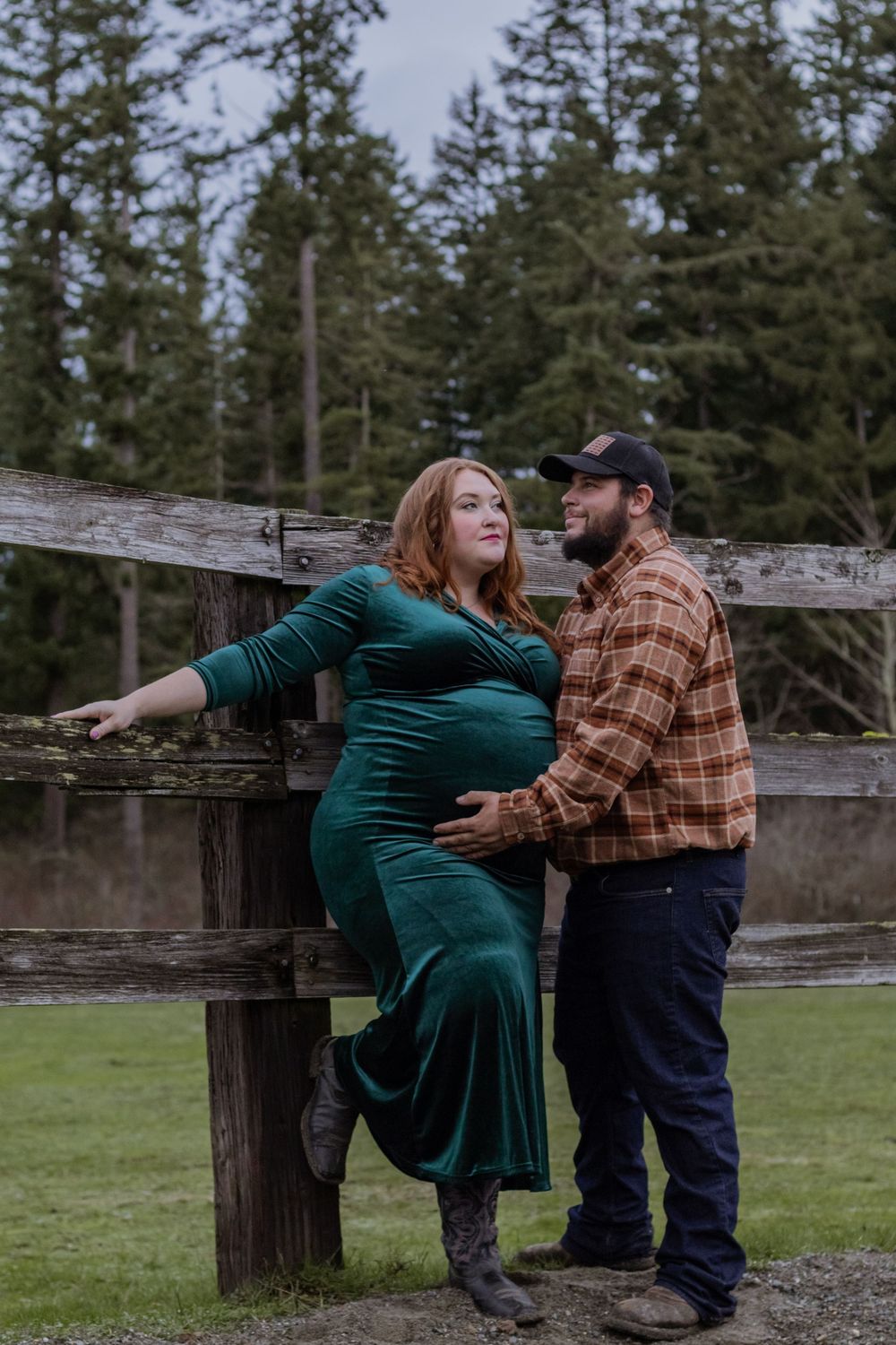 Pregnant woman in green velvet dress and partner leaning against rustic wooden fence outdoors.