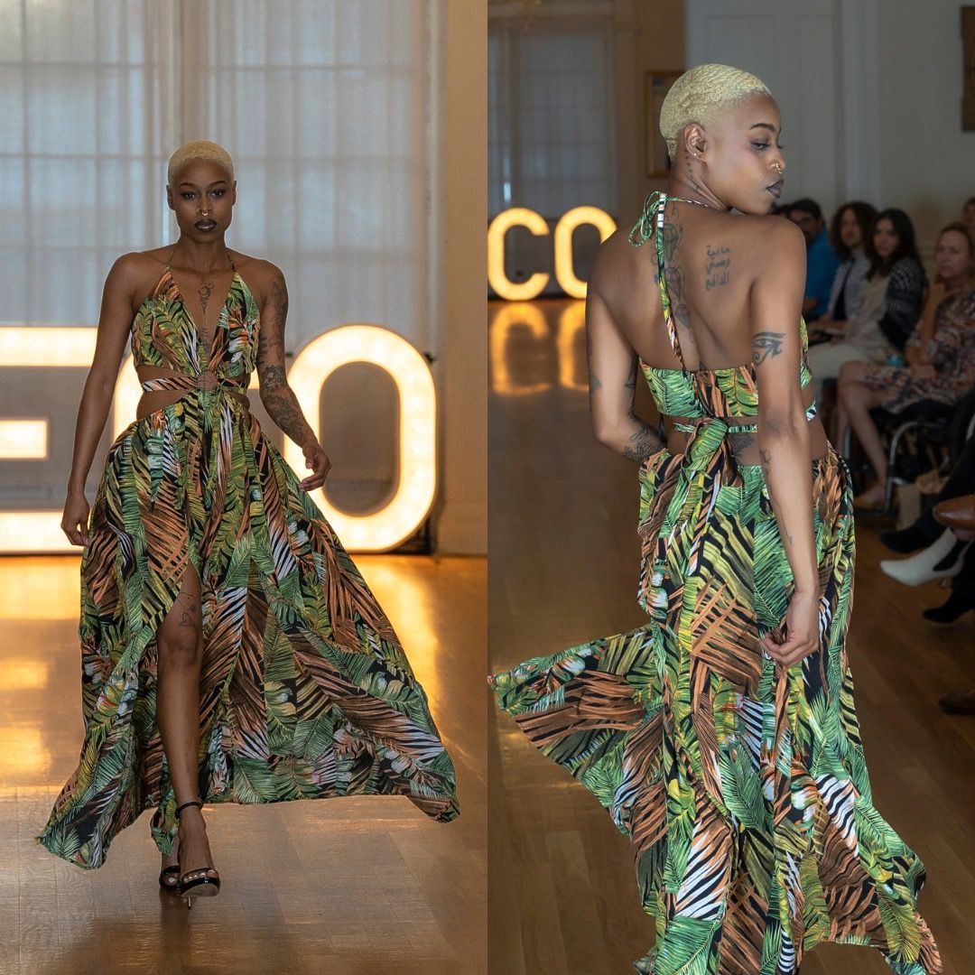 Model walks runway in flowing tropical print evening gown with dramatic side slit during fashion show.