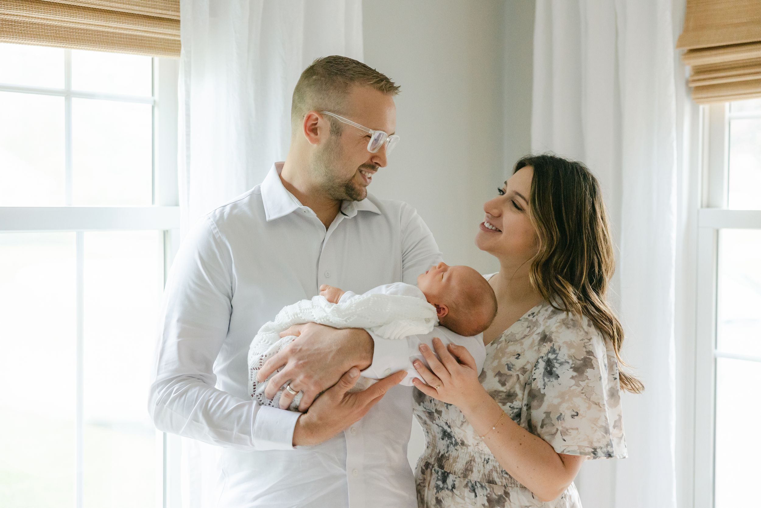 A couple stands by bright windows in their home, lovingly holding and gazing at their newborn baby.