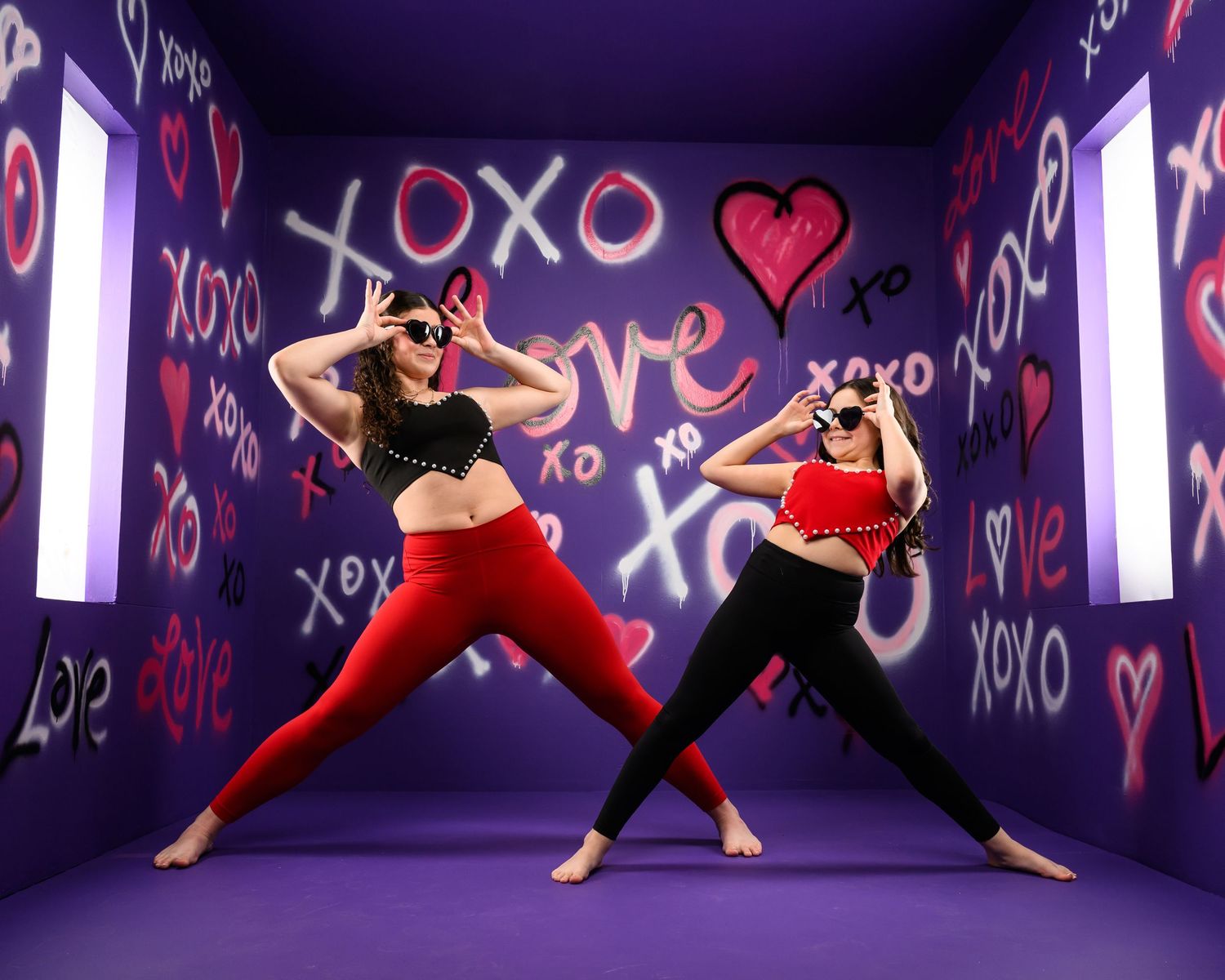 Dancers in black outfits strike dynamic poses against a purple wall decorated with white heart and XO graffiti designs.