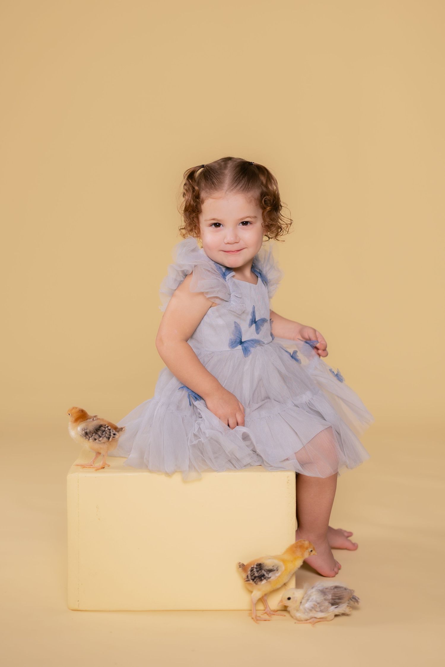 Baby in light blue tulle dress sits on cream-colored block with stuffed animal companions in studio setting.