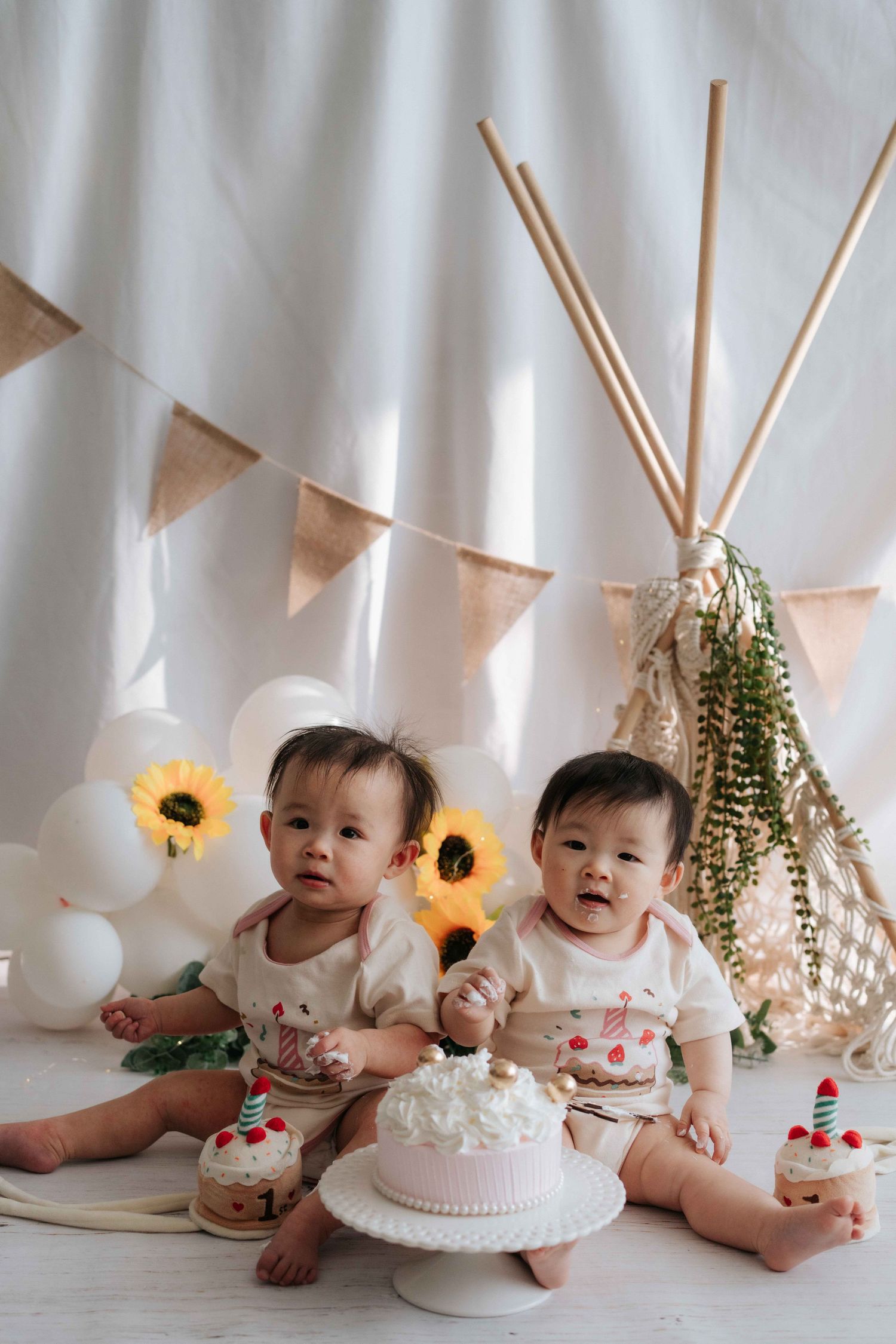 Baby cake smash photo session with white backdrop, sunflowers and pink cake with first birthday decorations.