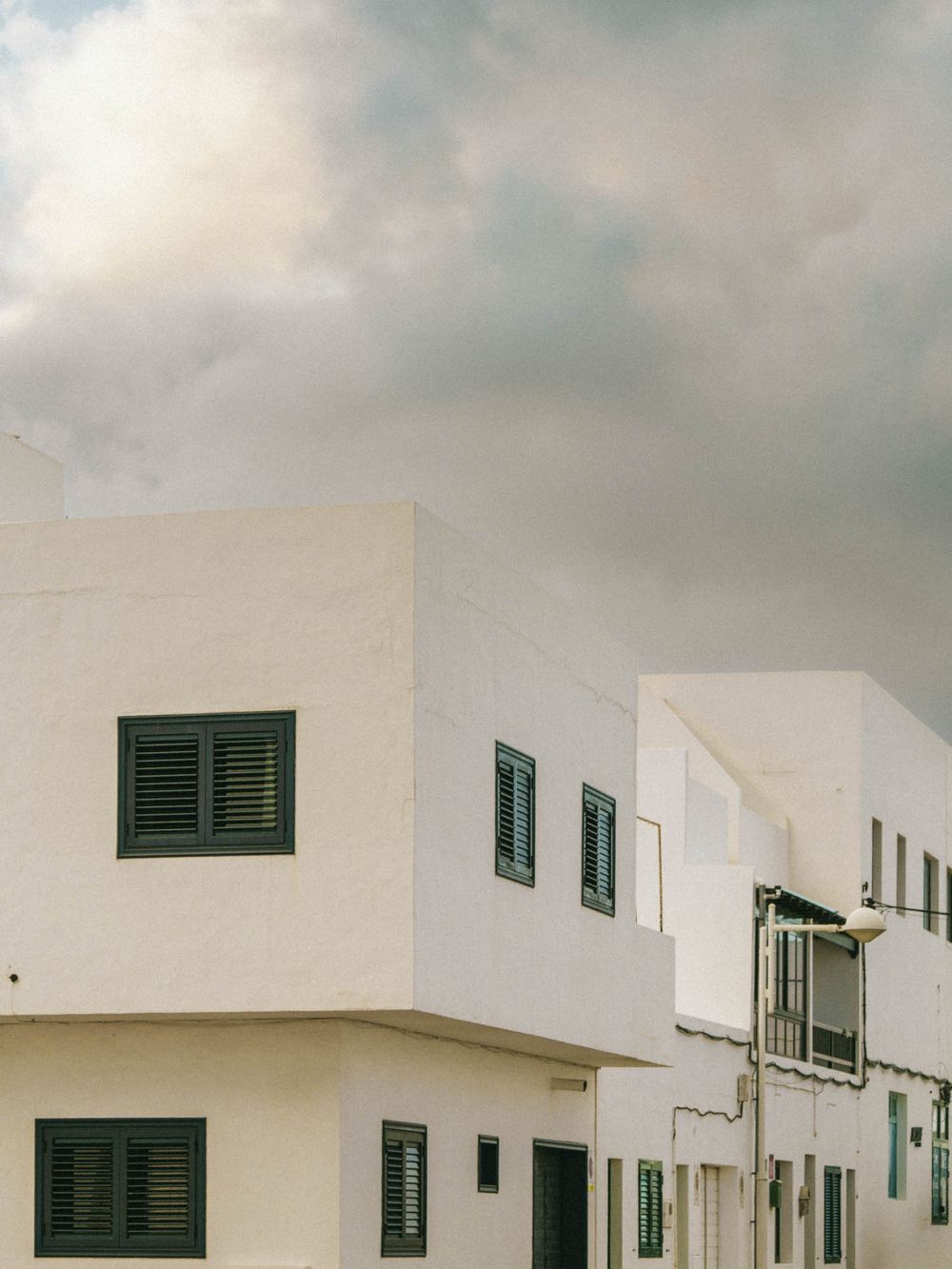 White Mediterranean style buildings with green shutters against a cloudy sky in a coastal village setting.