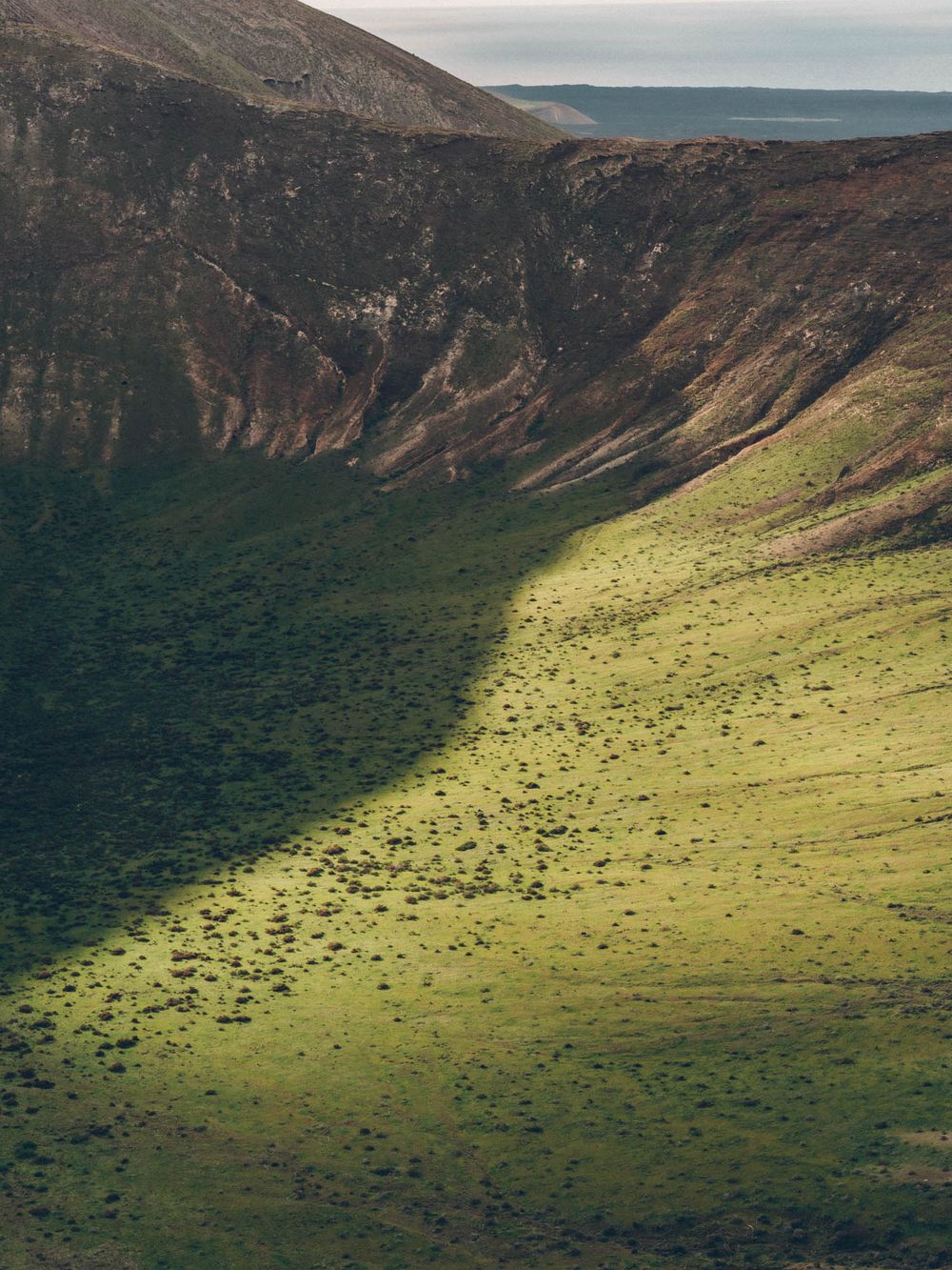 Aerial view of a vast volcanic crater with sunlit green slopes and dark rocky cliffs in Hawaii's natural landscape.