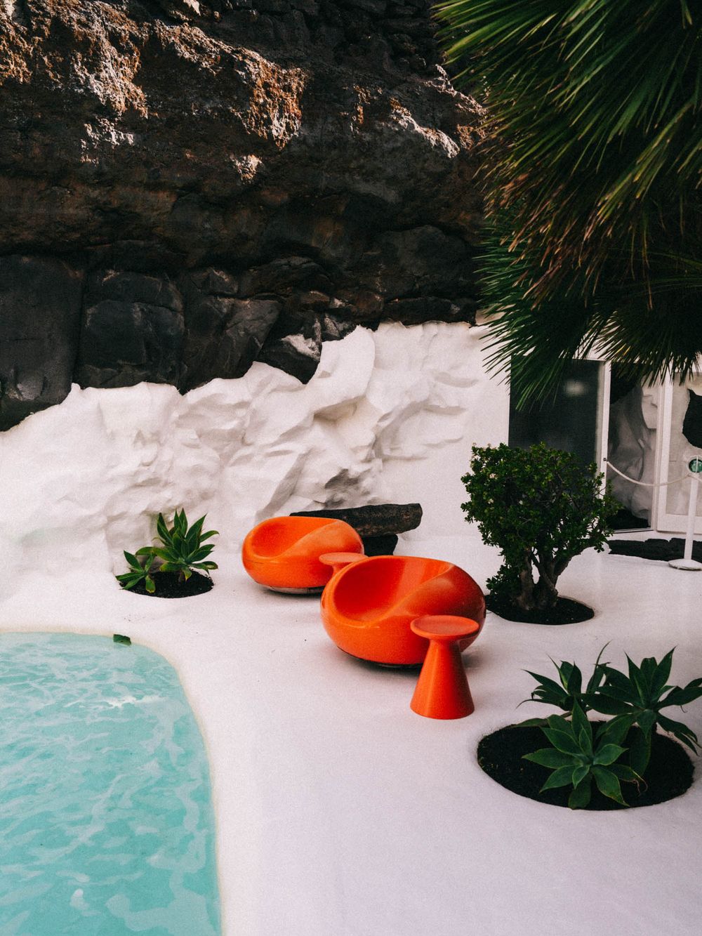 Modern orange chairs and tropical plants create a striking contrast against white snow and turquoise water in a cave setting.