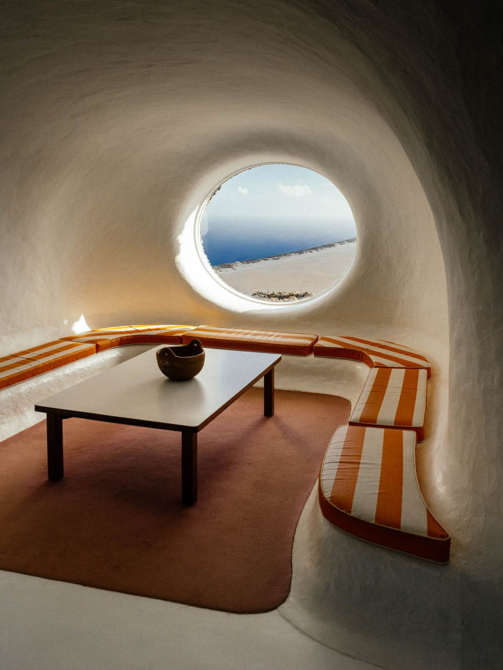 A minimalist room with curved white walls, built-in seating, and a round window overlooking the ocean.