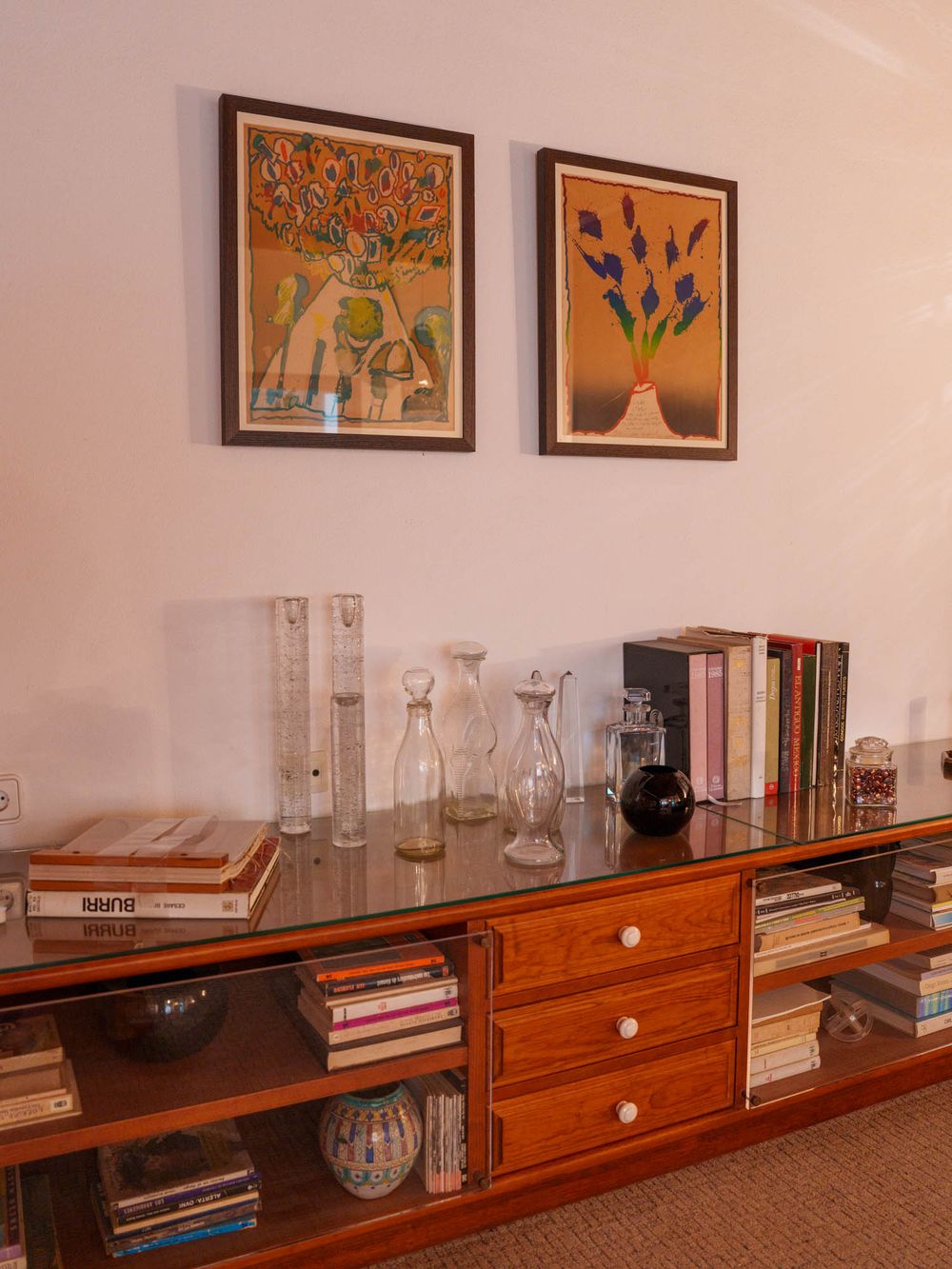 A long wooden shelf with glass vases and books displayed beneath colorful framed artwork on a beige wall.