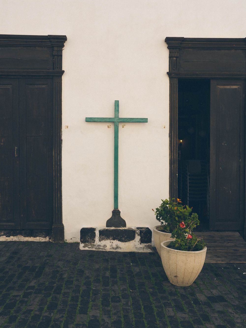 A green cross mounted on a white wall with dark wooden shutters and stone pavement creates a minimalist religious scene.