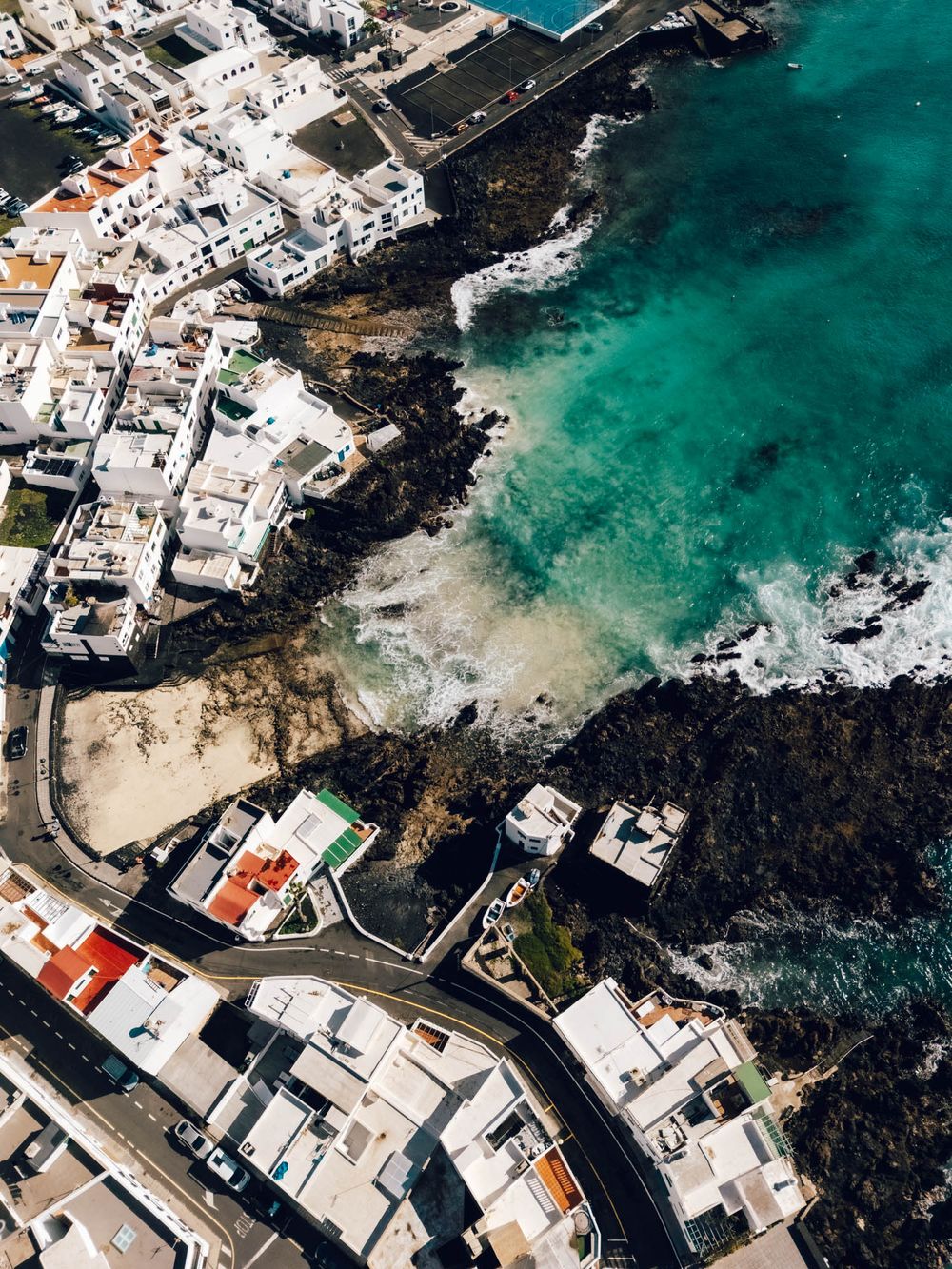 Aerial view of white coastal buildings and turquoise waters meeting volcanic black rocks in a Mediterranean seaside town.