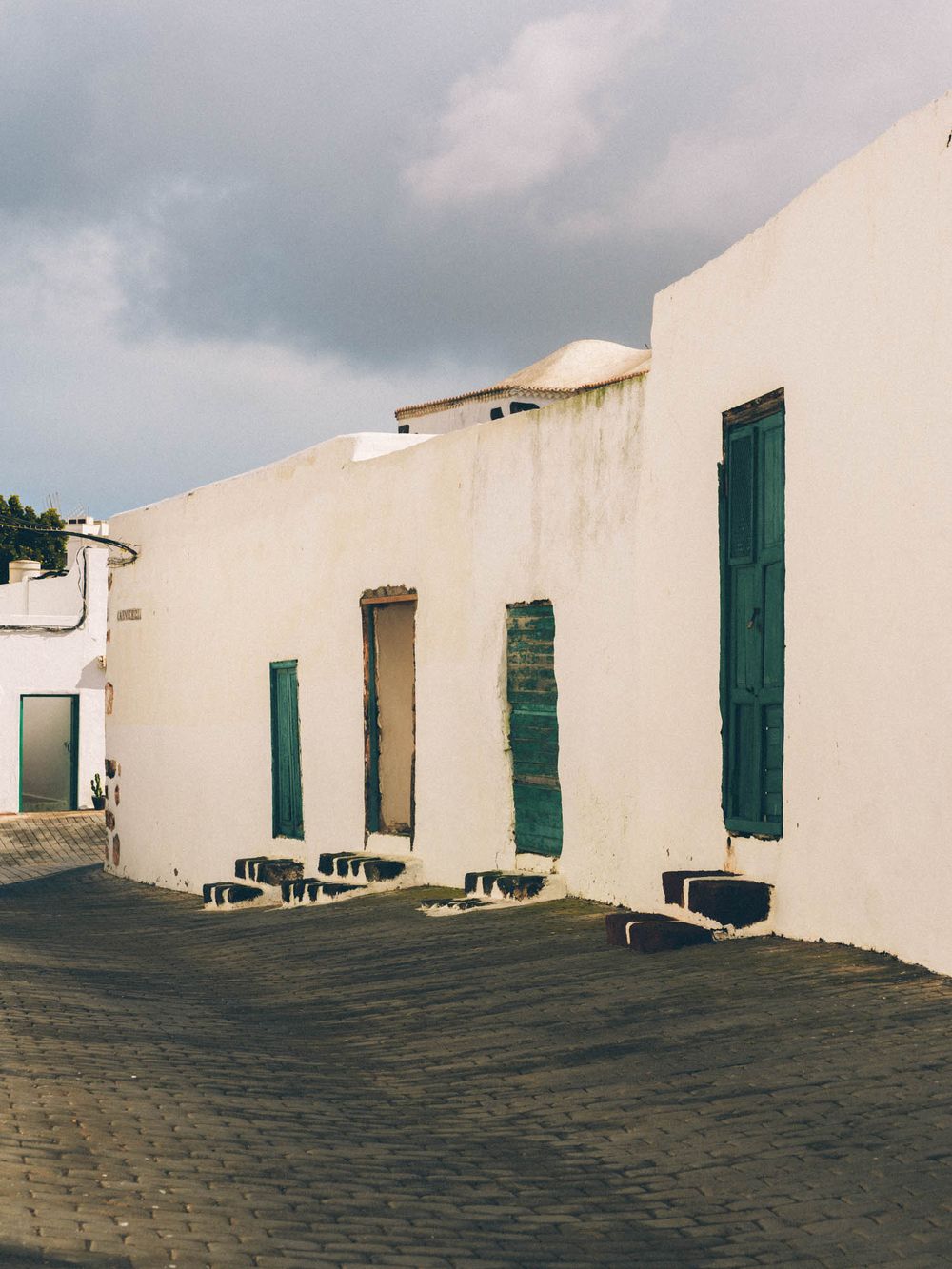 White stone buildings with green shutters line a cobblestone street against a cloudy sky in a historic Mediterranean town.