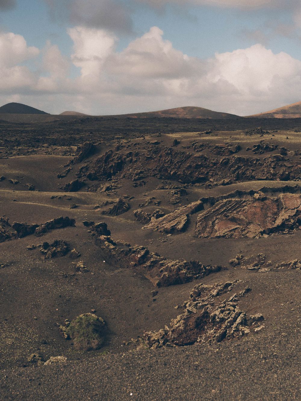 Panoramic view of a rugged volcanic landscape with dark rocky terrain and mountains under cloudy skies.