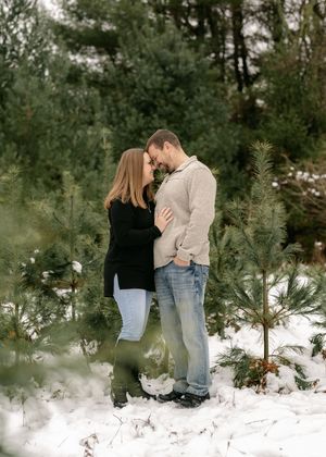 A couple stands together in a snowy winter forest setting with evergreen trees creating a romantic outdoor atmosphere.