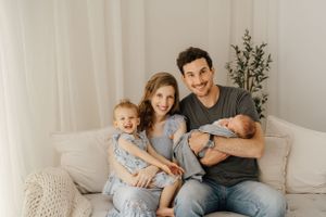 A family sits together on a cozy beige couch in a neutral-toned room with a decorative plant in the background.