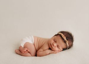 Sweet sleeping newborn in delicate headband poses curled up on soft cream backdrop in diaper.