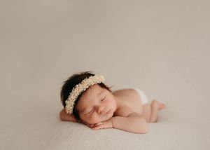 Sleeping newborn baby wearing delicate white lace headband rests peacefully on beige backdrop during professional photoshoot.