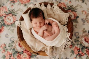 A peaceful newborn sleeps in a wooden bowl lined with soft cream fabric and lace on a floral patterned backdrop.