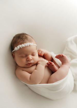 Peaceful newborn baby wearing a delicate white headband sleeps while wrapped in soft white fabric.