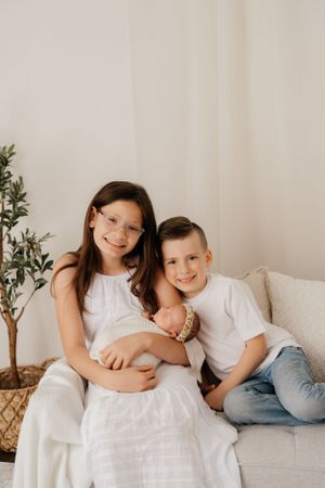 Two children sit together on a white couch with a small baby, next to a decorative indoor plant.