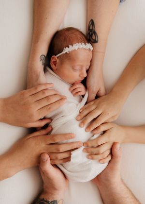 Newborn baby wrapped in white swaddle being gently held by multiple caring hands against light background.