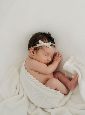 Sleeping newborn wearing white bow headband nestled in soft white textured blanket.