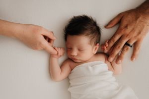 A sleeping newborn baby wrapped in white fabric with gentle hands touching their face and arm.