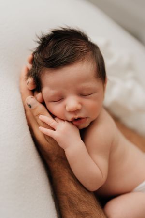 A sleeping newborn baby rests peacefully against a parent's supportive hand on white bedding.