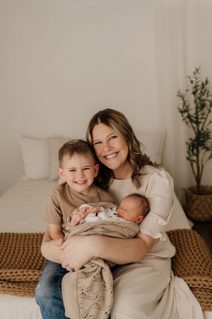A tender family portrait photoshoot in warm neutral tones showing siblings bonding with a newborn baby on a textured surface.