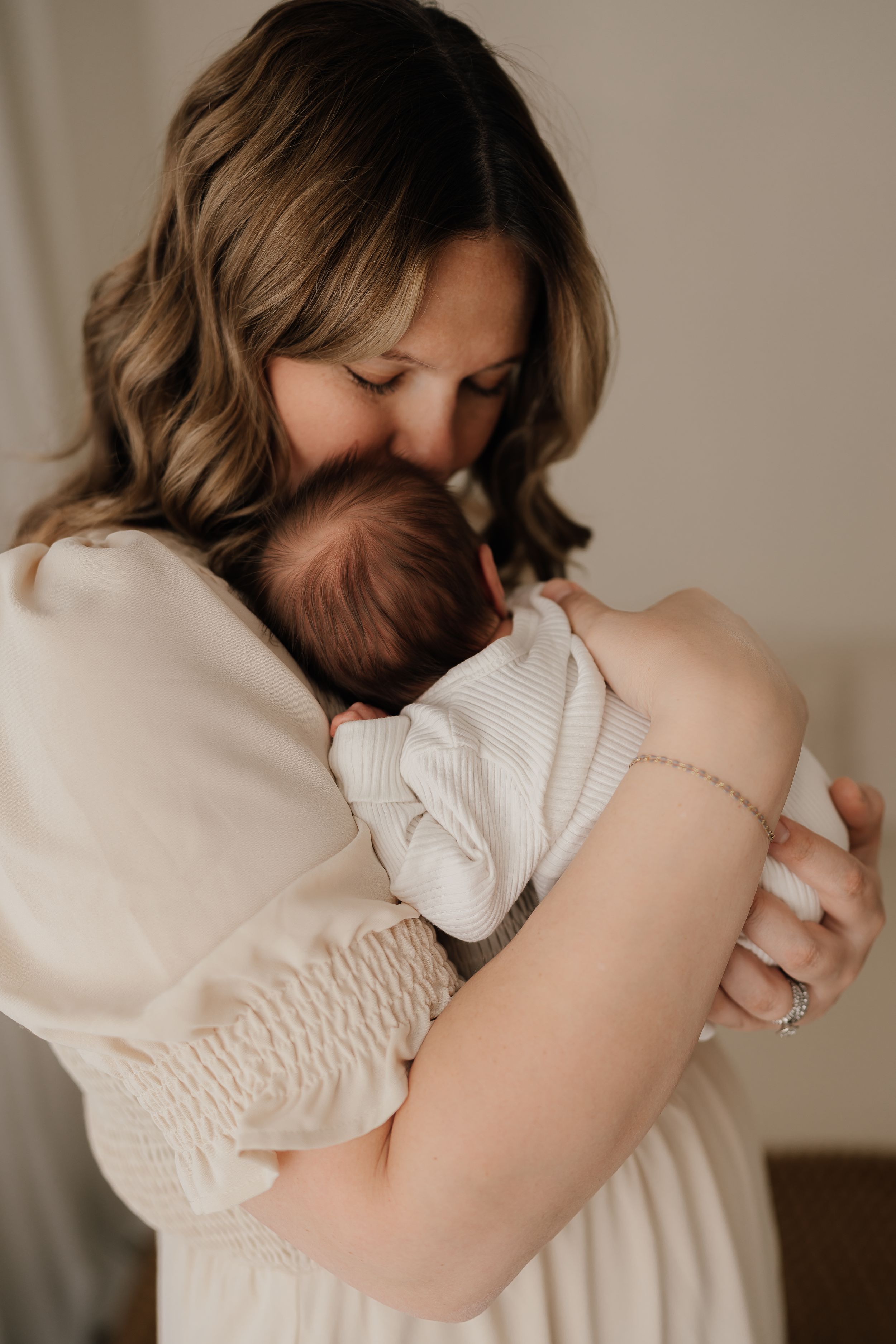 In a warm beige setting, a mother shares a tender embrace with her newborn baby against a soft neutral backdrop.