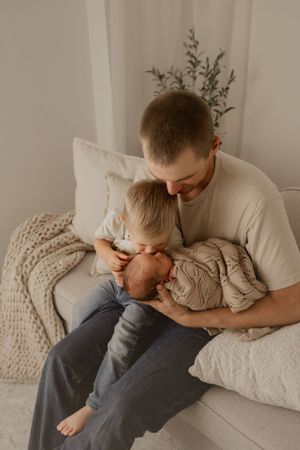 A tender family moment on a cozy bed with white pillows and a knit blanket in soft natural lighting.