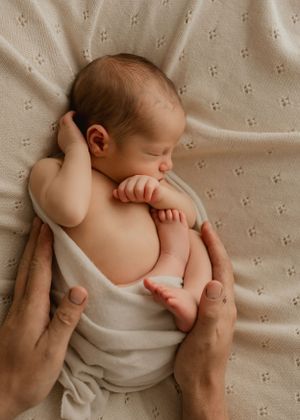 A peaceful sleeping newborn baby swaddled in beige fabric lies curled on a textured blanket with adult hands gently supporting.