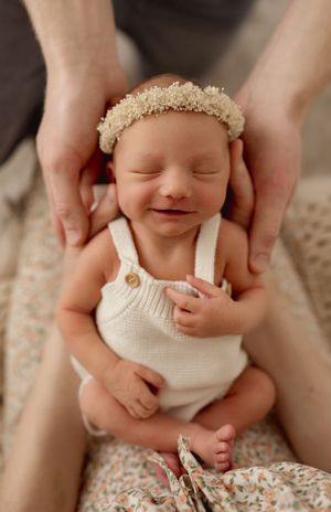 A peaceful newborn wearing a white knit romper and lace headband smiles while being cradled by gentle hands.