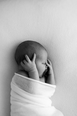 A peaceful black and white photo of a sleeping newborn wrapped in white cloth with hands near face.