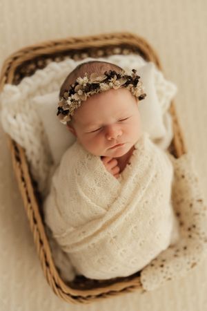 Sleeping newborn swaddled in cream wrap lying in wicker basket with floral headpiece on soft white blanket.