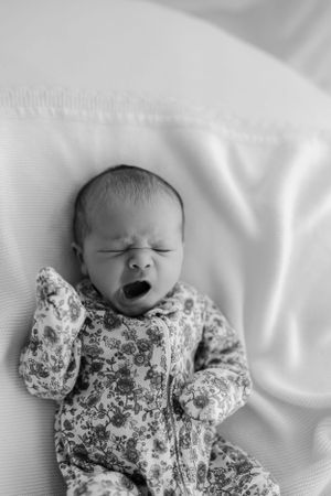 Black and white portrait of a sleeping newborn baby wearing a floral onesie on white bedding while yawning.