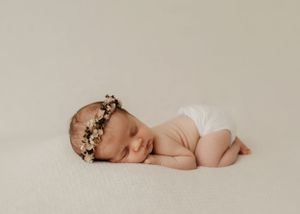 A newborn baby wearing a delicate floral crown sleeps peacefully on a cream-colored backdrop in a white diaper.