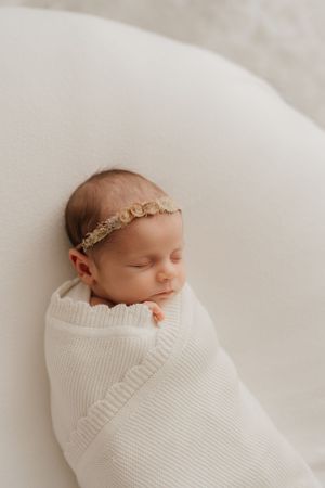 A sleeping newborn baby swaddled in a white knit blanket wearing a delicate gold headband on cream background.