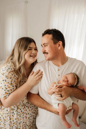 A couple shares a tender moment while holding their newborn baby in a bright, naturally lit room.