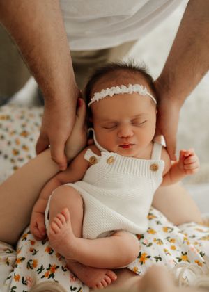 Sleeping baby wearing white knit romper and lace headband rests on floral print bedding while receiving gentle head massage.